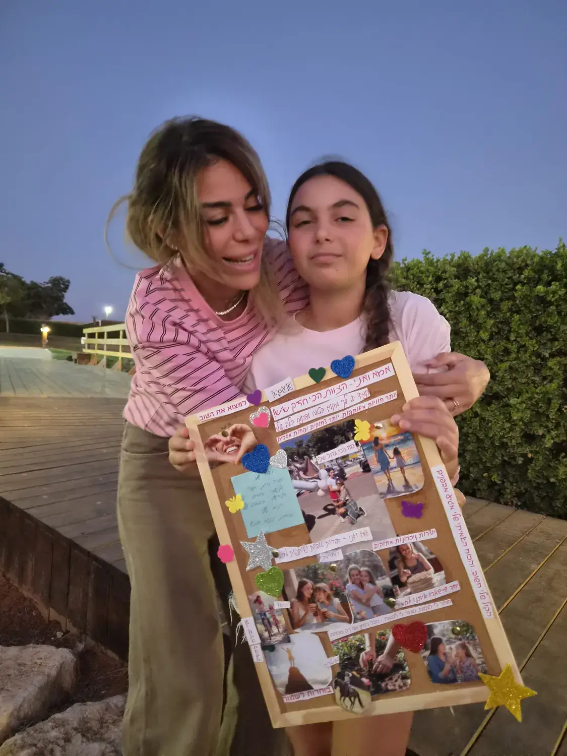 אמא מחבקת את הבת שלה מאחור, שתיהן מחזיקות לוח יצירה צבעוני